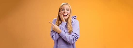 Amused excited speechless happy young blond woman 25s in hoodie cannot believe luck happiness pointing right amazed smiling broadly touch cheek surprised receive awesome unexpected gift photo