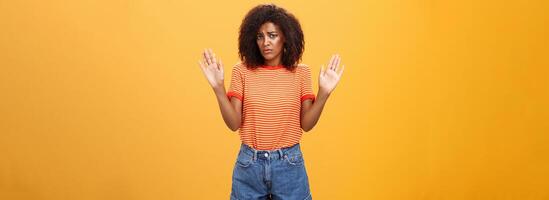 Silly insecure and sad dark-skinned female model in trendy striped t-shirt and shorts raising arms in surrender frowning being uninvolved and unaware standing clueless over orange background photo