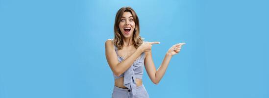 Unaltered shot of excited amused adult attractive woman with gapped teeth pointing left and smiling broadly from happiness and joy standing delighted and thrilled over blue background photo