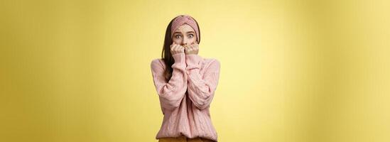 Stunned scared timid insecure cute young girl frightened gasping from fear pressing hands to mouth standing in stupor shocked, horrified stupified against yellow background being victim of abuse photo