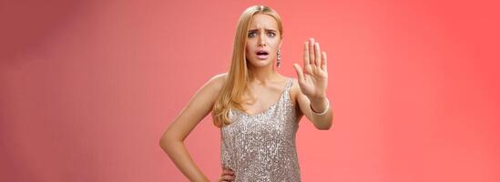 Freaked out displeased bothered insecure blond woman in silver glittering dress extend palm stop enough prohibition rejection gesture bothered pissed annoying clingy man nightclub, red background photo