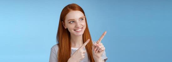 Attractive redhead female model pure clean skin ginger long hair pointing looking left intrigued smiling satisfied indicating interesting advertisement intrigued using new product, blue background photo