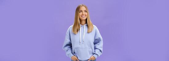 Winter comming. Dreamy kind and cute young girl with straight fair hair and tan wearing hoodie looking at upper left corner with nice white smile and admiring expression holding hand in pockets photo