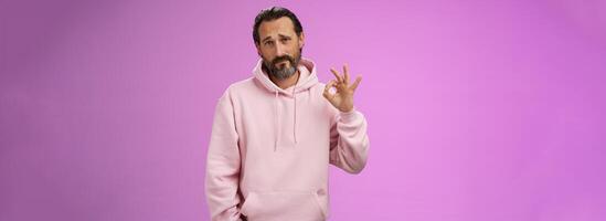 Not bad may better. Portrait handsome mature bearded father wearing trendy hoodie frowning awkward show okay ok normal gesture unimpressed but agree, standing purple background doubtful unsure photo