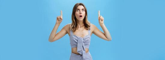 Waist-up shot of curious good-looking stylish modern woman dropping jaw saying wow while lifting head up looking and pointing upwards with amazed, surprised expression posing against blue background photo