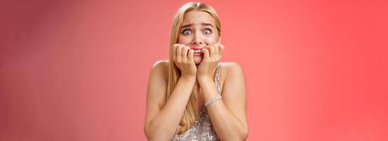 Frightened afraid panicking young cute blond woman biting nails pop eyes scared camera look terrified stunned speechless stare forward standing red background gasping trembling fear photo