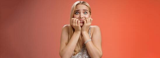 Frightened afraid panicking young cute blond woman biting nails pop eyes scared camera look terrified stunned speechless stare forward standing red background gasping trembling fear photo