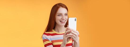 Redhead girl having fun recording hilarious friend actions hold smartphone look display amused shooting funny telephone standing orange background satisfied smiling delighted photo