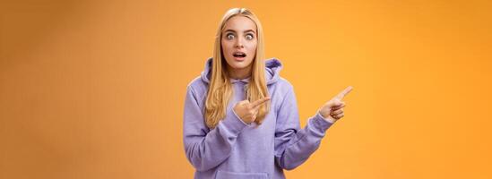 Wow astonished impressed good-looking blond european female in hoodie pop eyes wondered surprised questioned drop jaw pointing left excited asking if gossip true, standing orange background photo