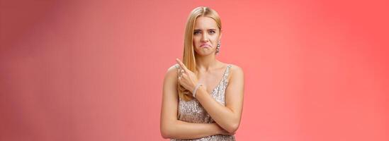 Jealous displeased angry young revengeful blond ex-girlfriend in luxurious silver shiny dress frowning pouting pointing upper right corner displeased pissed standing fed up upset red background photo