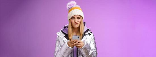 Perplexed displeased cute blond stylish girl unwilling anser message cringing reluctant frowning holding smartphone receive disappointed perplexing text, standing unsure purple background photo