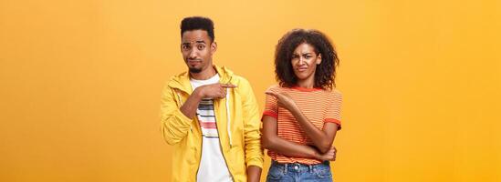 Friends getting shocked someone thought they are dating. Displeased mocking african american female frowning and grimacing from aversion while pointing at guy who indicating at girl over orange wall photo