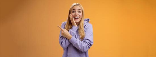 Amused excited speechless happy young blond woman 25s in hoodie cannot believe luck happiness pointing right amazed smiling broadly touch cheek surprised receive awesome unexpected gift photo