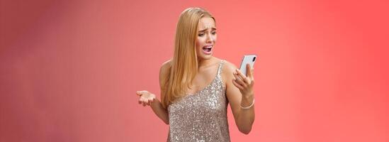Troubled concerned arrogant young blond woman complaining yelling smartphone cannot call friend no signal holding smartphone look mobile display pissed moody arguging, red background photo