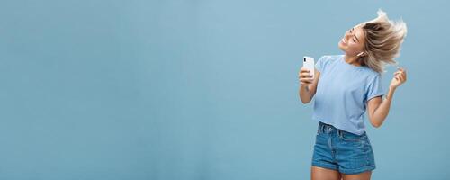 Girl enjoying cool bits in brand new wireless earphones advertising earbuds in own blog recodring via smartphone dancing from joy and delight smiling listening music over blue background photo