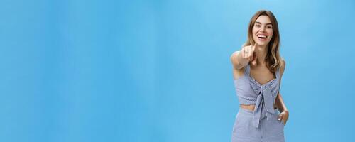 Haha your are funny. Portrait of amused carefree attractive young female in matching outfit pointing at camera as if having fun over photographer laughing out loud being in good mood over blue wall photo