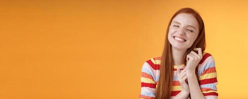 Happy dreamy romantic young tender ginger girl fantasizing creating love story imagination smiling broadly delighted close eyes touching hair strands recalling nice memory, standing orange background photo