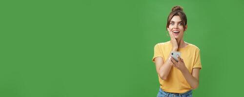 Surprised touched and impressed beautiful european girl with diastema smiling broadly touching cheek from amazement and joy holding smartphone looking happy at camera after reading cool news photo