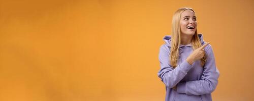 Amused carefree joyful blond european woman in hoodie pointing looking right pleased smiling happily enjoying interesting fascinating peromance making choice in store shopping, orange background photo