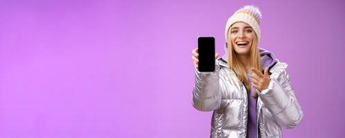 Satisfied amused good-looking blond girl suggest take look smartphone display smiling happily pointing mobile phone delighted talking about awesome new app features, standing purple background photo