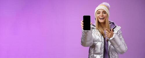 Satisfied amused good-looking blond girl suggest take look smartphone display smiling happily pointing mobile phone delighted talking about awesome new app features, standing purple background photo