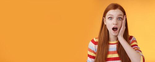 Shocked impressed concerned ginger girl drop jaw gasping stunned slap cheek wide eyes surprised hear disturbing terrible news standing orange background sympathizing awful story photo