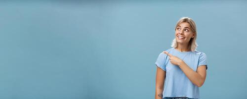 Waist-up shot of creative and dreamy happy attractive blonde female in casual t-shirt looking and pointing at upper left corner with admiration and joy standing over blue background amused and curious photo
