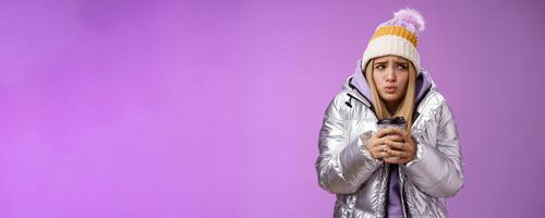 Trembling uncomfortable cute pouting young blond girl feel freezing cold winter snowy weather outside shaking low temperature hold take-away hot coffee cup warming up, purple background photo
