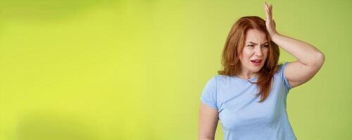 Something important slip my mind. Concerned worried upset redhead mature woman punch forehead turn away frustrated frowning disappointed forget cancel appointment stand green background photo