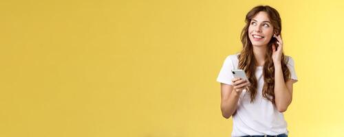 Dreamy happy cheerful curly-haired girl look around contemplate beautiful summer weather listening music touch wireless earbud calling friend talking via earphones hold smartphone yellow background photo