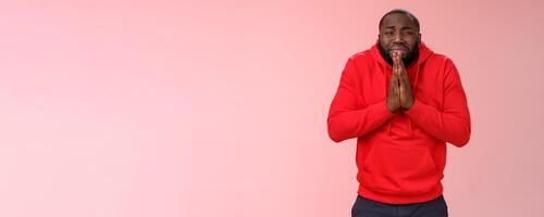 Miserable african american man beard in red hoodie stooping make supplicating upset grimace grying sobbing asking help say please hold hands pray begging mercy, apologizing need money photo