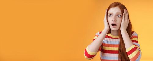 Worry insecure redhead european panicking girl grab head hands both sides gasping open mouth shocked stare frightened upset watching terrible accident standing concerned orange background photo