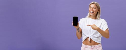 Satisfied happy beautiful tanned woman with blond hairstyle and tattoos holding smartphone showing device screen and pointing at gadget with index finger smiling at camera over purple wall photo