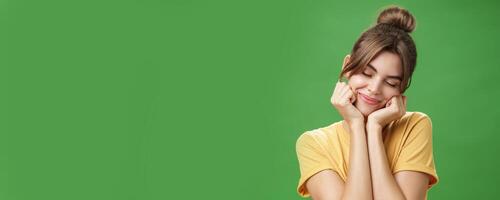 Tender sensual timid european girl in yellow t-shirt leaning head on shoulder touching cheeks with hands closing eyes and smiling with soft grin feeling nostalgic and romantic over green background photo