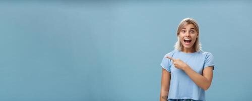 Hurry up and look. Excited and thrilled impressed charming blond female student in casual t-shirt smiling and yelling from amazement and happiness while pointing left staring surprised at camera photo