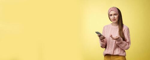 Puzzled confused upset questioned young cute girl in headband knitted sweater frowning clueless raising hand in dismay holding smartphone, cannot understand what happened cellphone over yellow wall photo