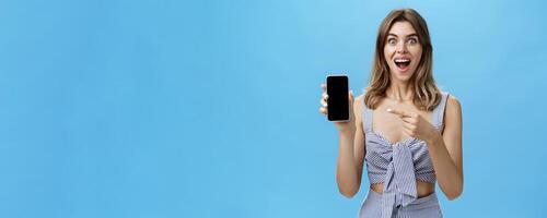 Excited happy woman with gapped teeth finally bought brand new smartphone holding device in hand pointing at cellphone screen showing cool app smiling broadly from joy against blue wall photo