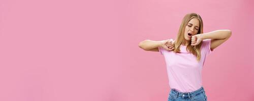 Too tired to work today. Lazy and exhausted attractive young female student doing homework all night yawning with closed eyes while stretching hands from after tiresome project over pink wall photo