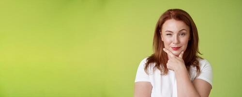 Hmm interesting choice. Intrigued cunning redhead middle-aged female hold hand face-line rub chin thoughtful smirking pleased raise eyebrow curious pondering alluring decision green background photo