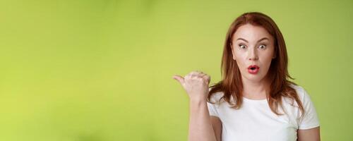 Close-up surprised amused redhead middle-aged housewife folding lips say wow impressed stare camera questioned ambushed pointing left thumb astonished wondering if promo true green background photo