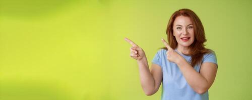 Curious enthusiastic gorgeous redhead female intrigued pointing upper left corner asking question interesting product talk shop assistant consulting promo stand green background blue t-shirt photo