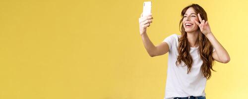 Positive charisamtic happy young girl curly-haired raise smartphone taking selfie show peace victory sign having call stand yellow background smiling mobile front camera posing lively photo