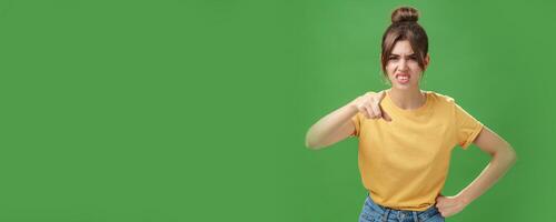 Portrait of arrogant impolite young woman in yellow t-shirt pointing at camera squinting and grimacing from disdain and scorn standing unimpressed judging against green background photo