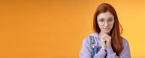 Silly guilty young shy redhead girlfriend asking forgiveness supplicating lower head look from under forehead frowning begging apology favour standing insecure sad pleading help, orange background photo