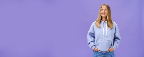 Winter comming. Dreamy kind and cute young girl with straight fair hair and tan wearing hoodie looking at upper left corner with nice white smile and admiring expression holding hand in pockets photo