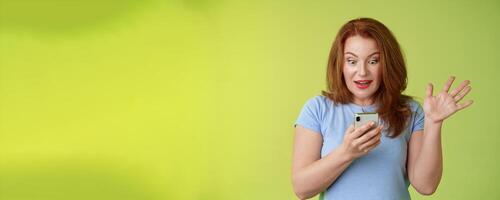 Surprised enthusiastic happy redhead mature middle-aged woman receive excellent message read perfect news social media hold smartphone stare astonished pleased mobile phone screen raise hand triumph photo