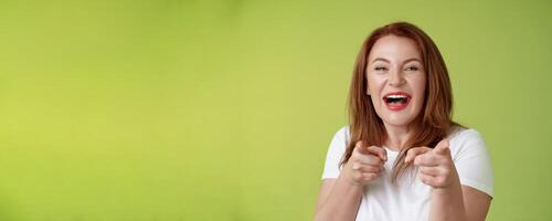You did best. Friendly joyful enthusiastic redhead ginger middle-aged female pointing index fingers camera finger pistol gesture smiling broadly congratulate cheer coworker stand green background photo