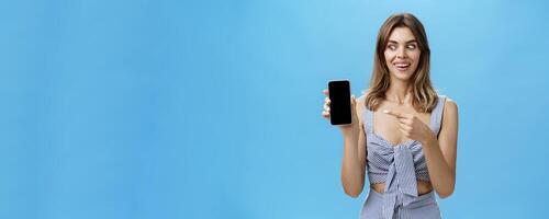 Portrait of charismatic astonished and happy woman with cute gapped teeth smiling axcited and surprised holding awesome smartphone pointing at device screen and looking at gadget amazed and delighted photo