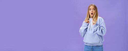 Woman scared of sudden loud noise in dark gasping looking shocked and concerned tilting backwards with frightened look pressing clenched fists to body from fear, wearing warm hoodie over purple wall photo
