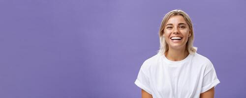 Close-up shot of joyful charming blonde female with delighted and pleased smile standing in white t-shirt over purple background spending time in awesome amusing company photo